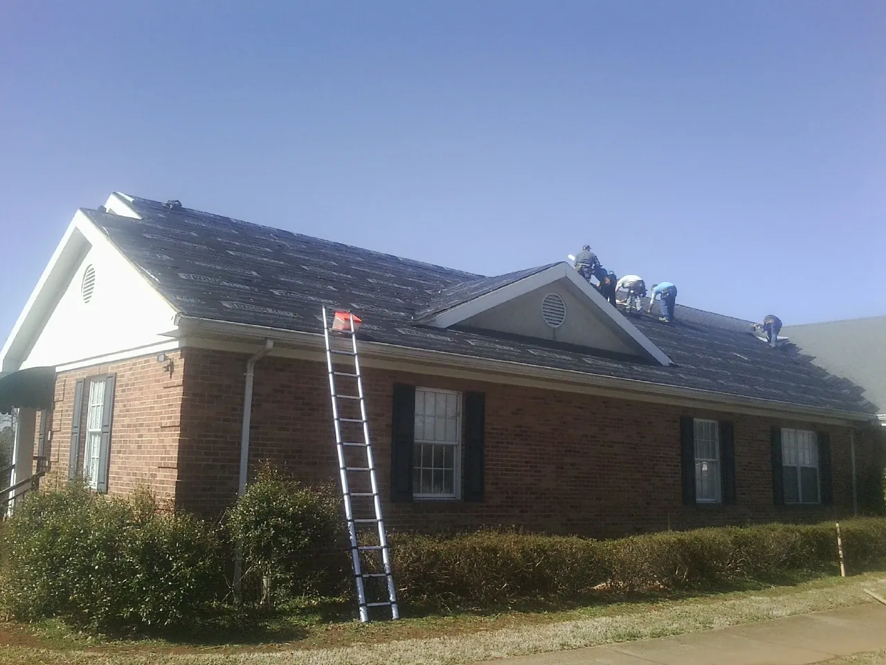 Crew tearing shingles off a brick ranch roof with a ladder set — Metal Roof Repair work by our crew in Wetumpka