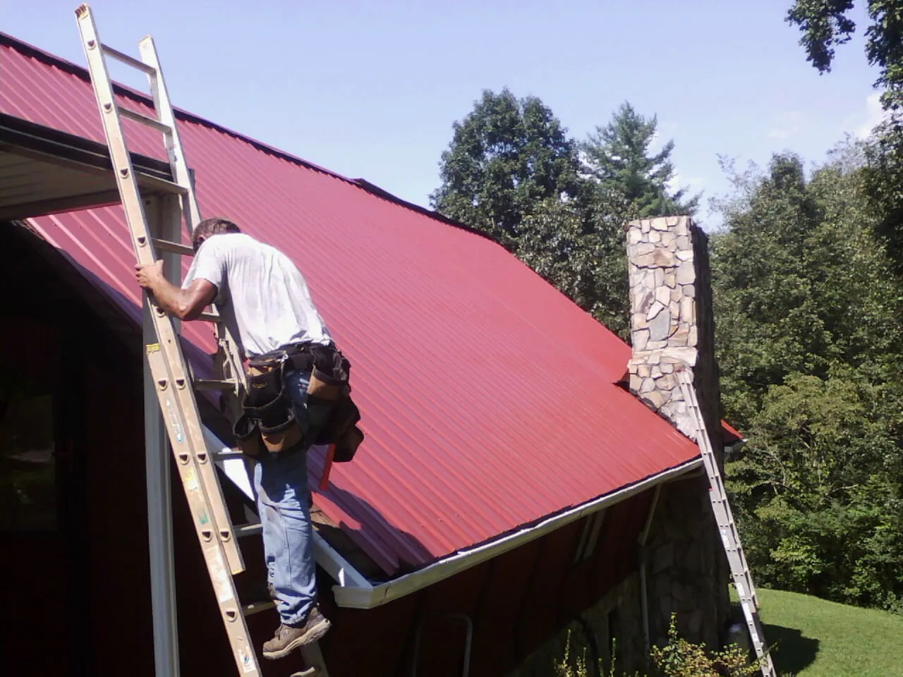 Roofer on a ladder beside a red metal roof and stone chimney — Soffit & Fascia Repair by Roofing Brothers in Wetumpka